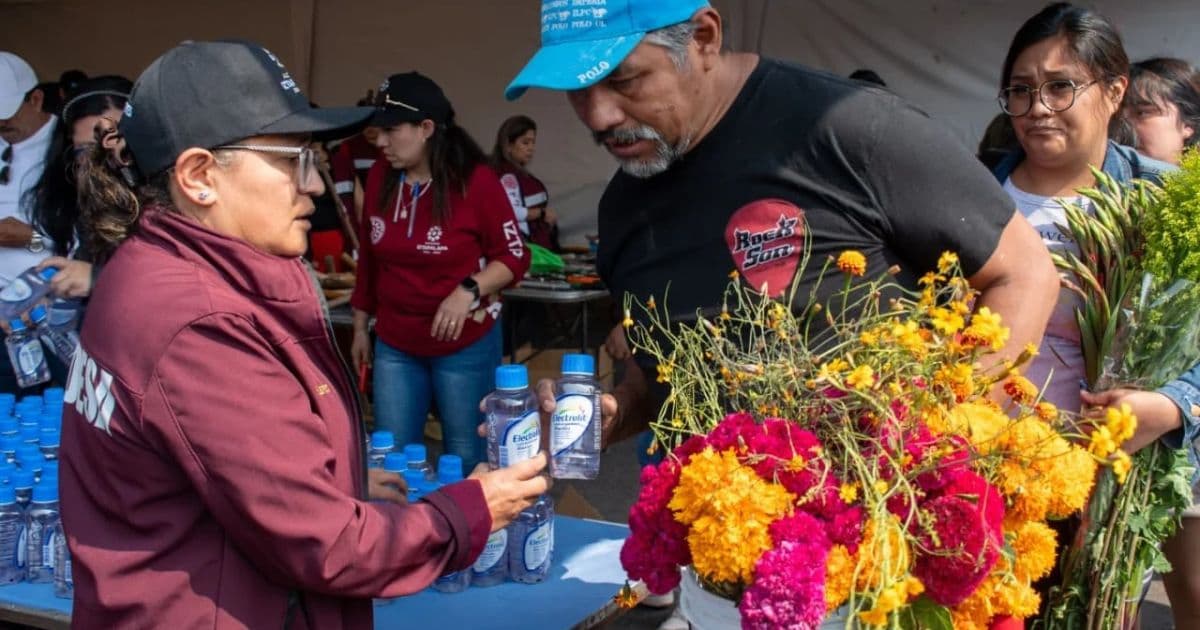 Una mujer entrega botellas de agua a un hombre que sostiene flores de cempasúchil, junto a una mesa de hidratación.