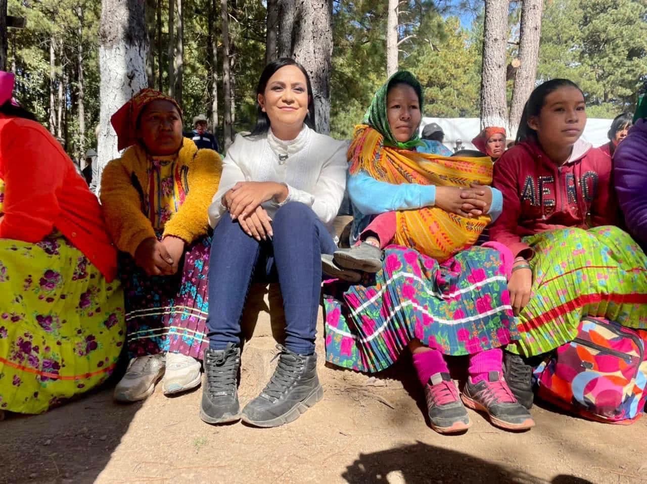 Ariadna Montiel sentada junto a mujeres y una menor de edad en un espacio arbolado, con vestimenta de colores y textiles tradicionales
