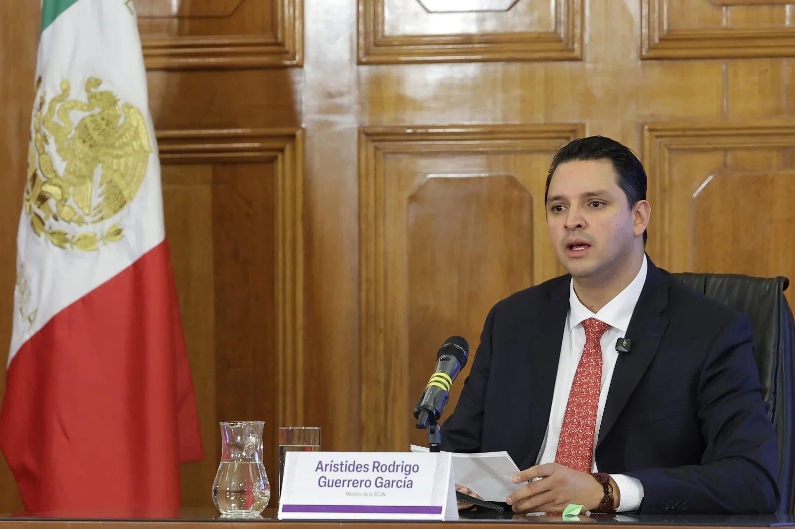 Arístides Rodrigo Guerrero García interviene ante un micrófono junto a la bandera de México