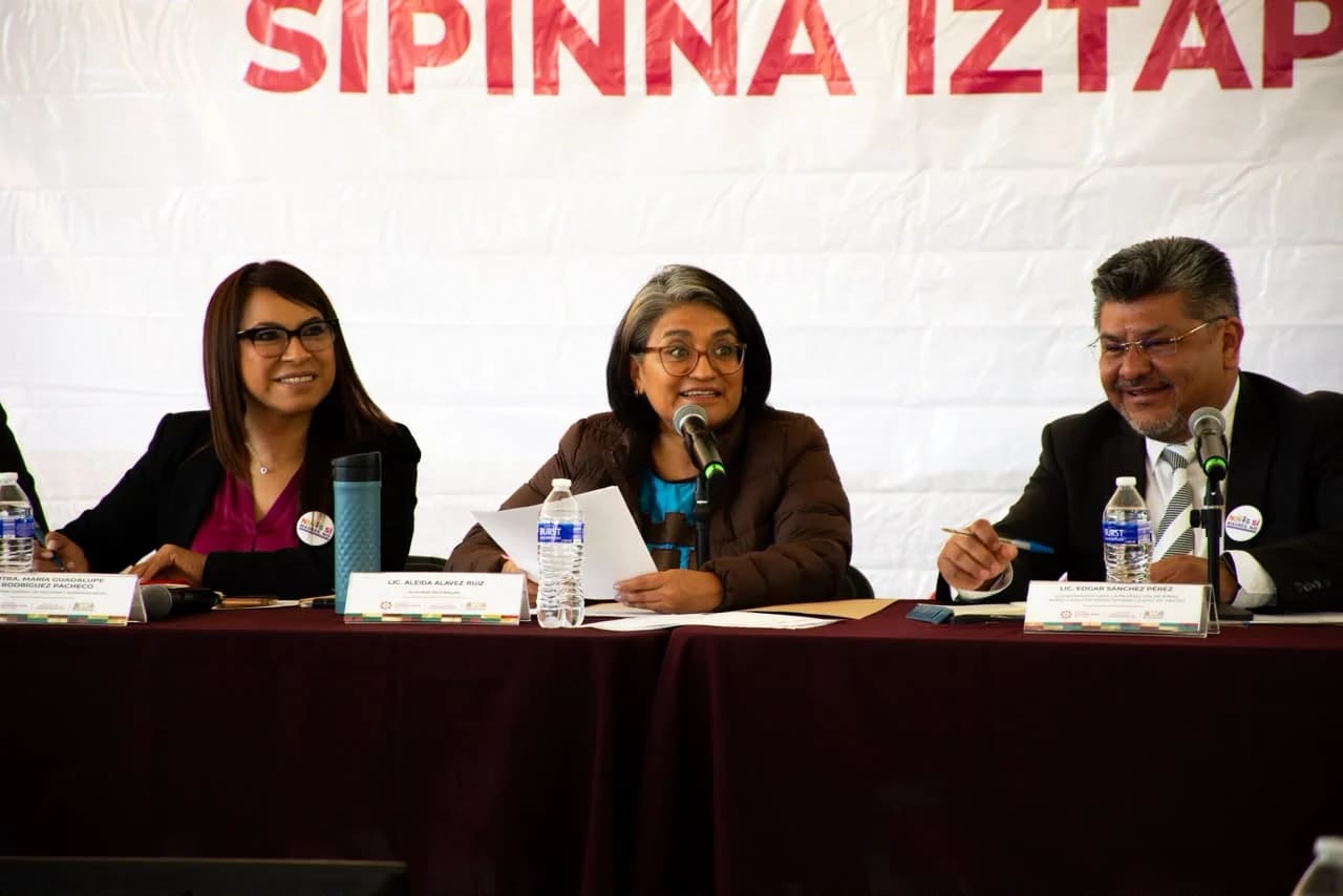Aleida Alavez Ruiz habla ante un micrófono durante una reunión de SIPINNA Iztapalapa, junto a otras dos personas en la mesa