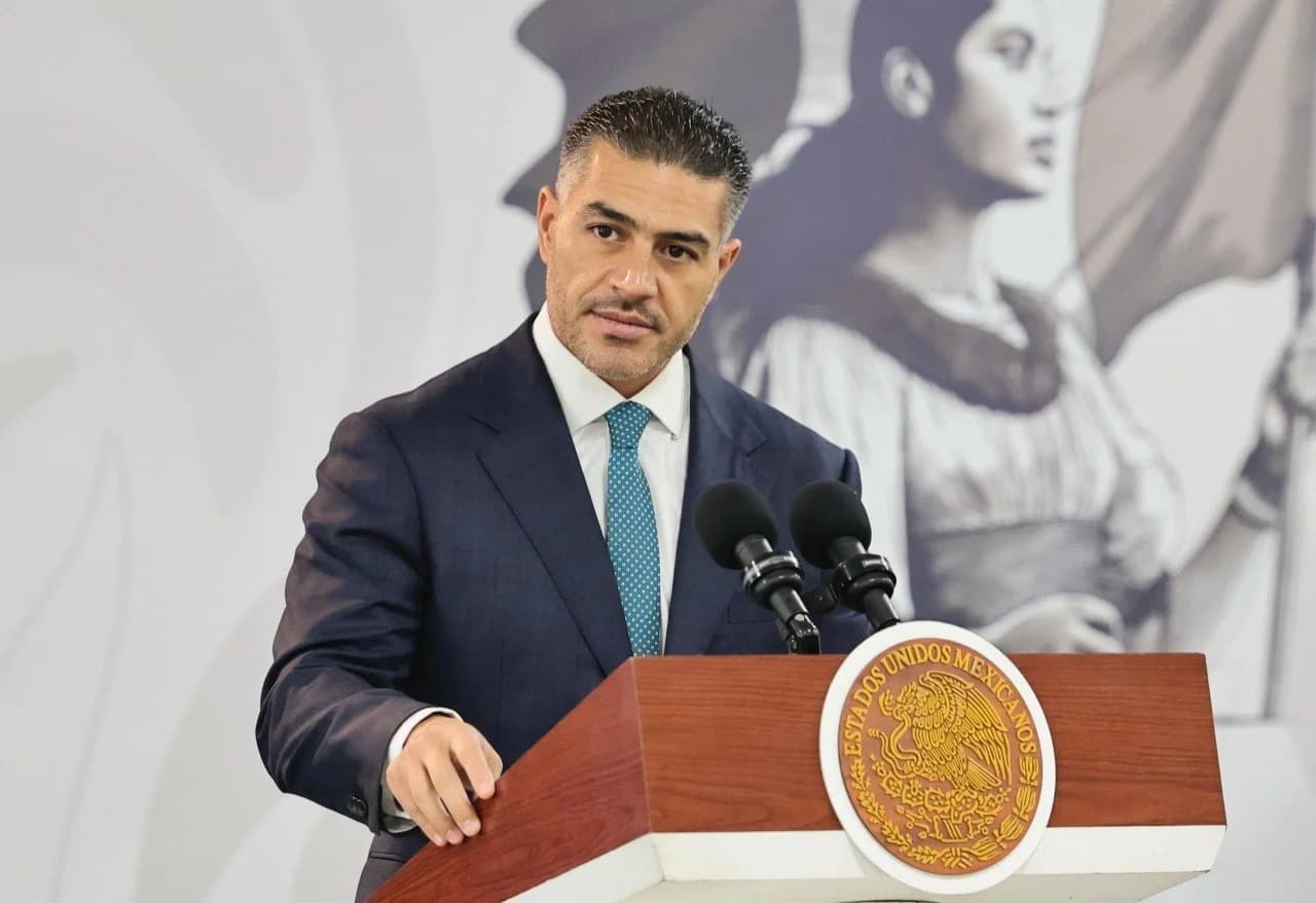 Omar García Harfuch, de traje azul marino y corbata turquesa, frente a un podio con el escudo nacional y dos micrófonos