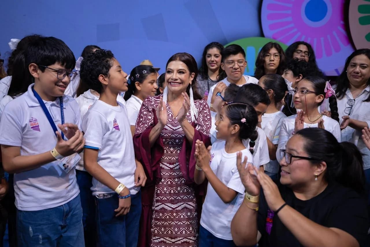 Clara Brugada sonríe y aplaude rodeada de niñas, niños y personas adultas durante un acto público.