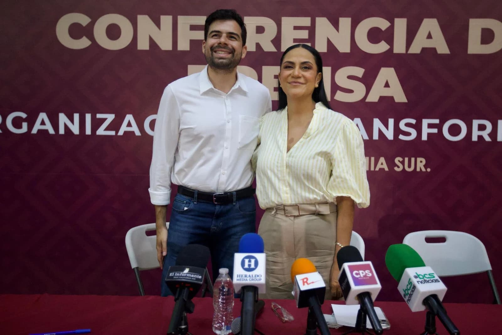 Dos personas posan juntas ante un fondo guinda de una conferencia de prensa, con micrófonos frente a ellas.
