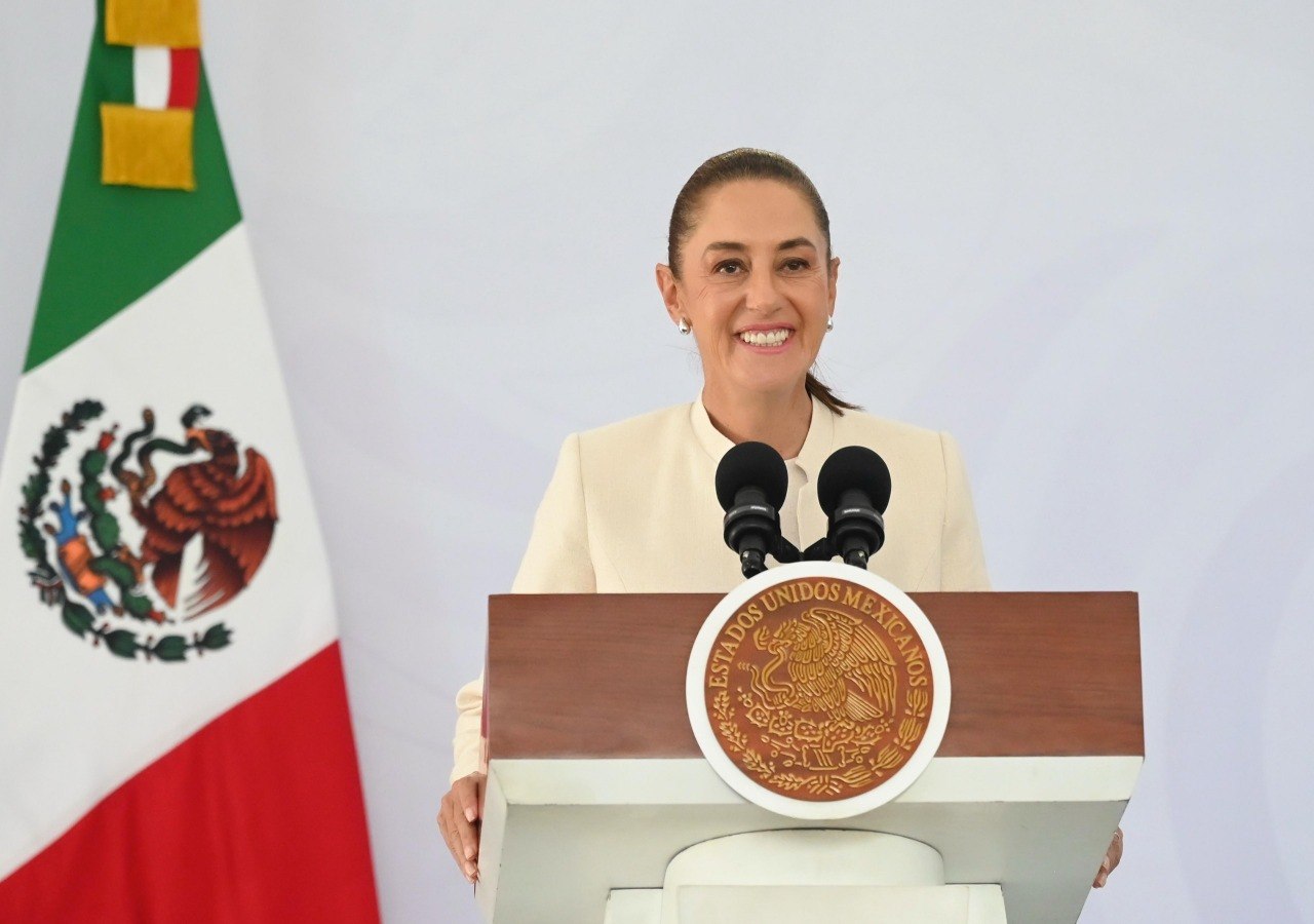 Una mujer habla desde un podio con el Escudo Nacional y la bandera de México a un costado.