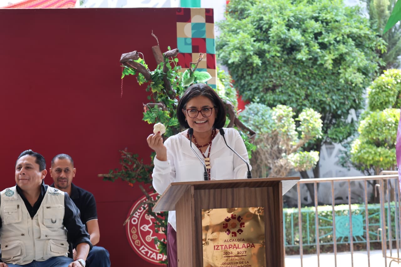 Una mujer habla desde un podio durante un evento de la Alcaldía Iztapalapa, con dos hombres sentados a un costado y vegetación al fondo.