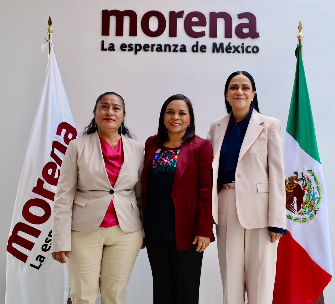 Tres mujeres posan juntas frente al emblema de Morena, entre una bandera del partido y la bandera de México.