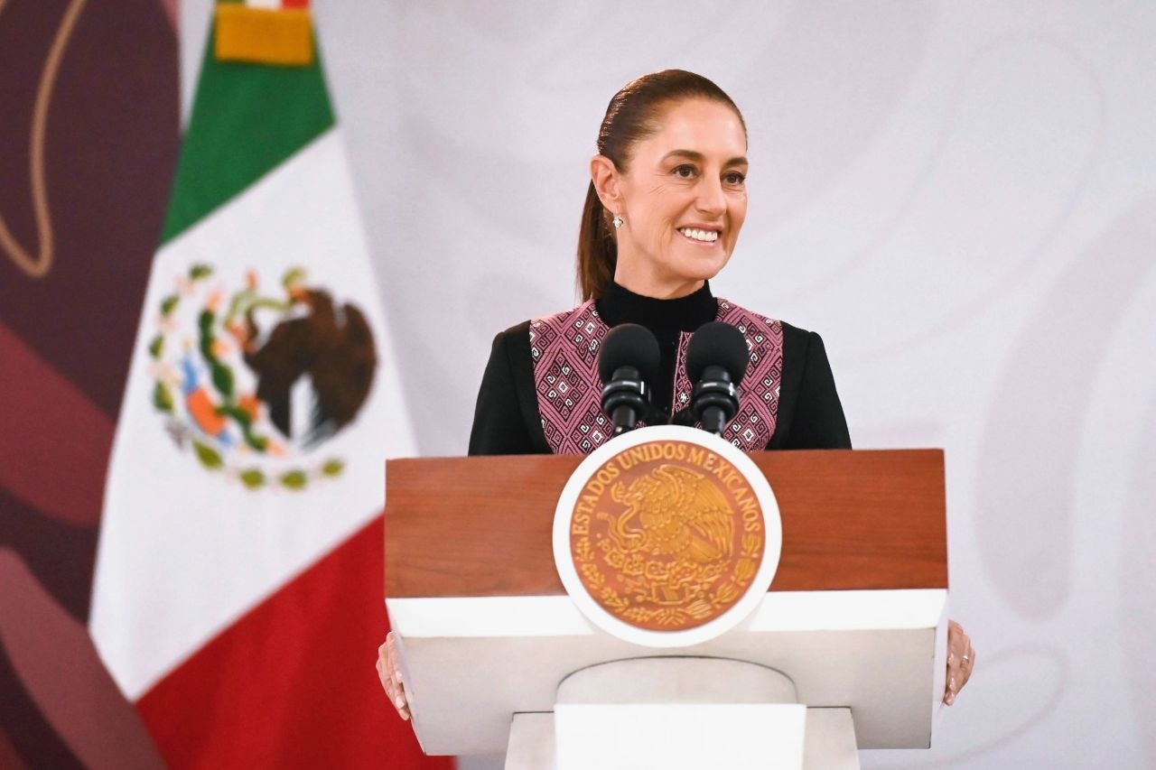 Presidenta de México frente al podio durante una conferencia, con la bandera nacional al fondo