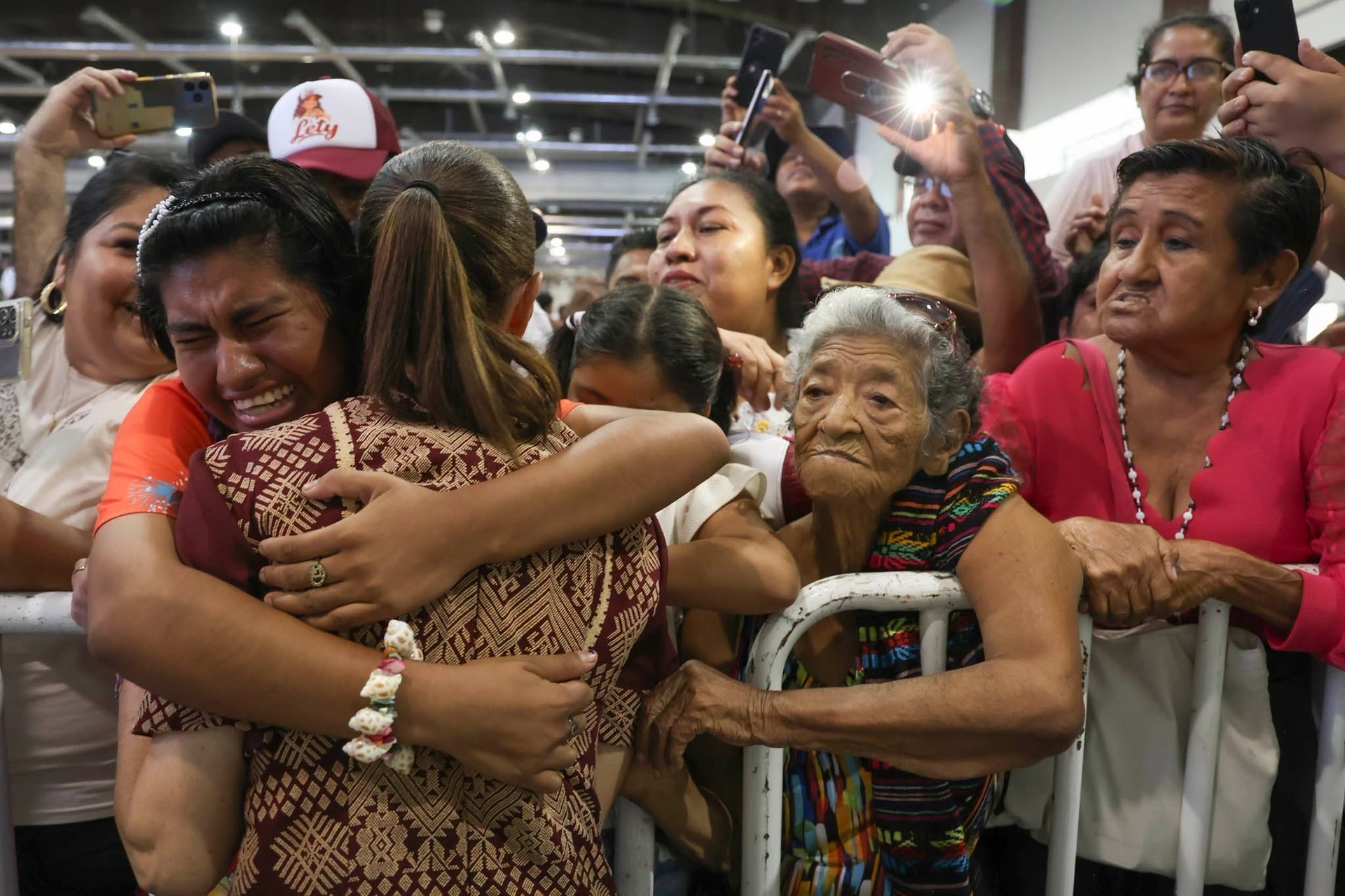 Claudia Sheinbaum abraza a una mujer entre asistentes a un acto público, mientras varias personas observan y toman fotografías.