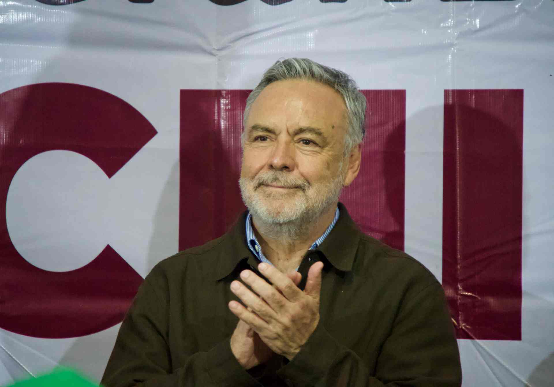 Hombre aplaude frente a un fondo con letras grandes en color guinda durante un acto público