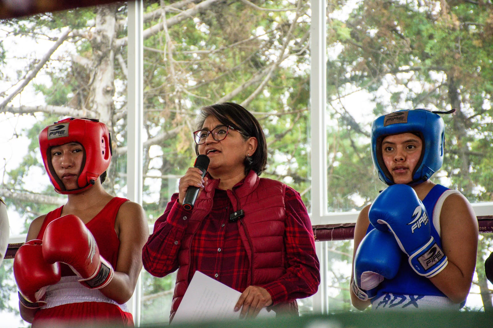 Una mujer habla con micrófono entre dos boxeadoras equipadas para competir.