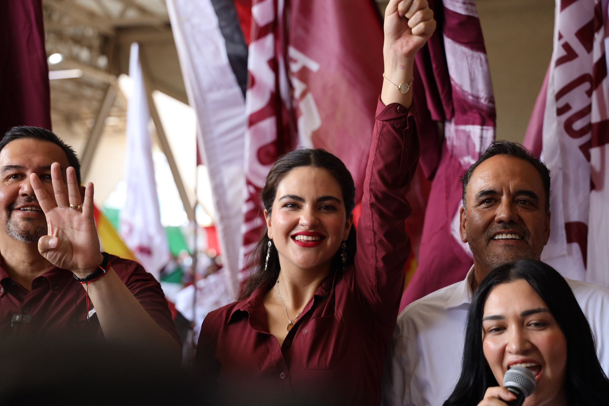 Mujer levanta el brazo durante un acto político con banderas de Morena al fondo