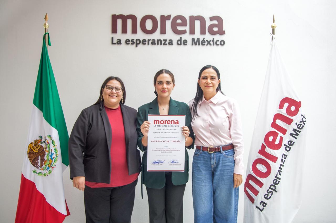 Tres mujeres posan frente a los emblemas de Morena y las banderas de México y del partido; la mujer del centro sostiene un documento