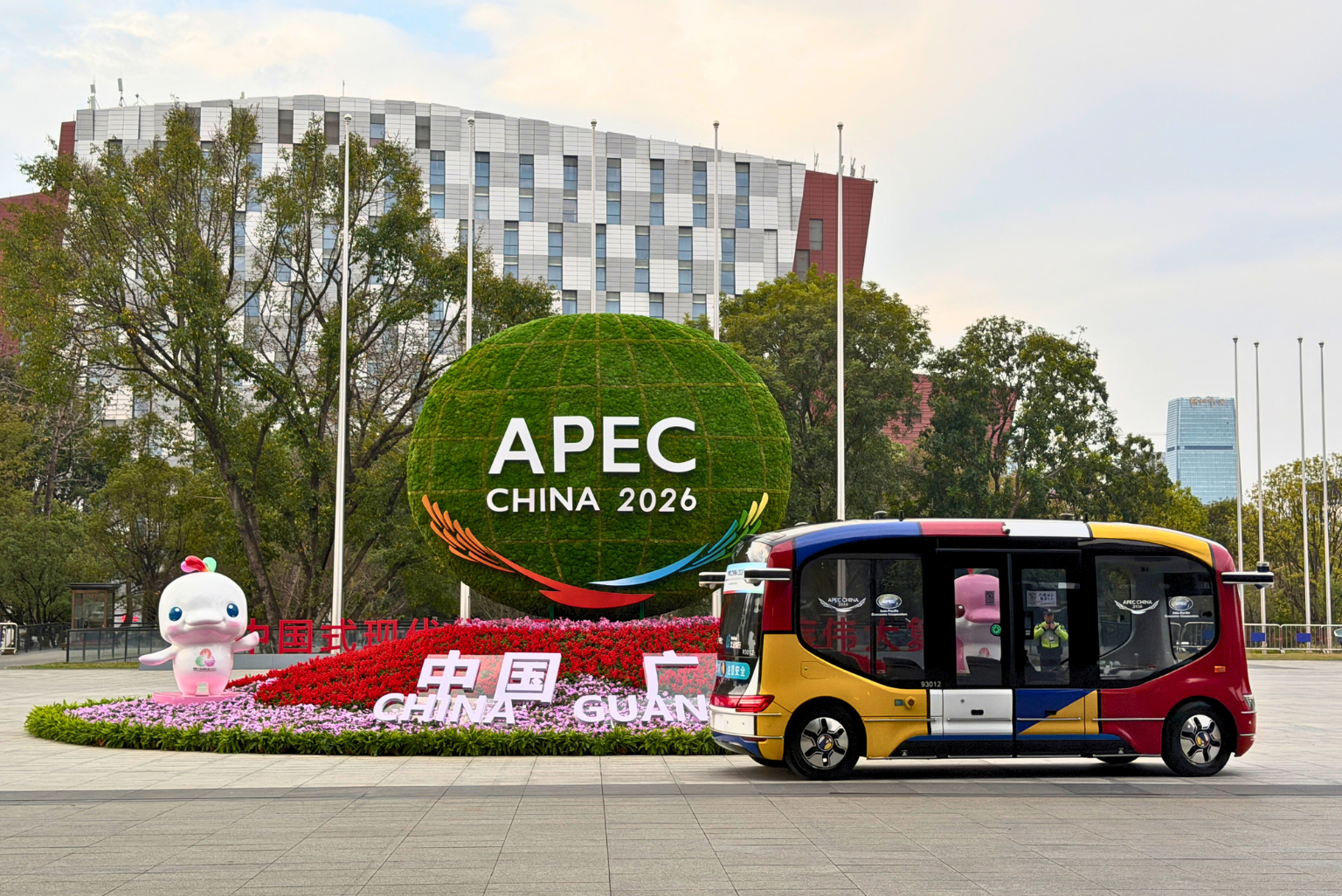 Instalación urbana con el letrero APEC China 2026, flores y un vehículo autónomo frente a edificios y árboles