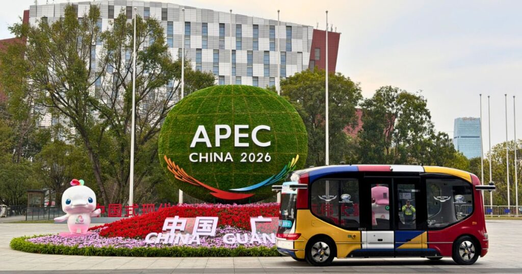 Instalación urbana con el letrero APEC China 2026, flores y un vehículo autónomo frente a edificios y árboles