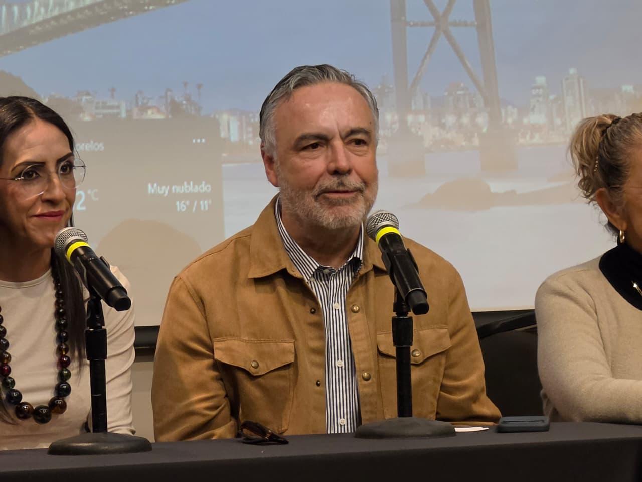 Hombre de cabello gris con chaqueta café habla ante un micrófono en una mesa de conferencia junto a otras participantes