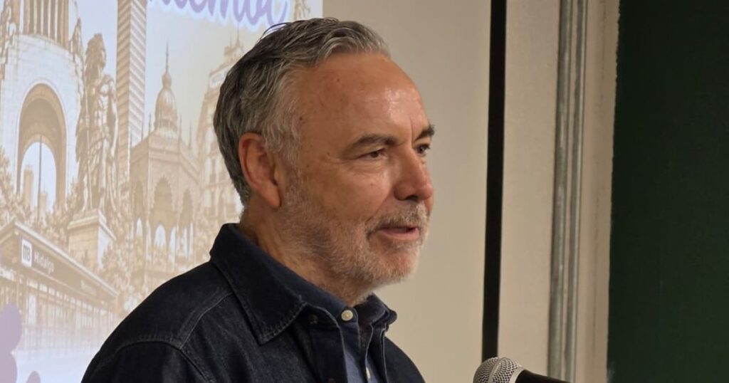 Hombre de cabello canoso participa ante un micrófono con una proyección de edificios detrás