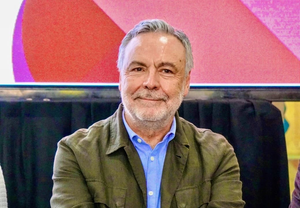 Hombre de cabello canoso y barba, con camisa azul y saco verde, sentado frente a una pantalla de colores