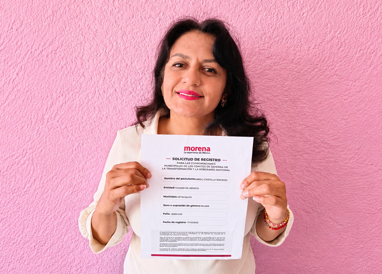 Mujer con blusa clara sostiene frente a una pared rosa una solicitud de registro con el logotipo de Morena.