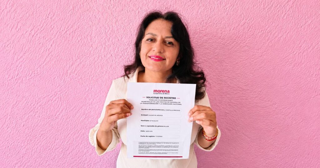 Mujer con blusa clara sostiene frente a una pared rosa una solicitud de registro con el logotipo de Morena.