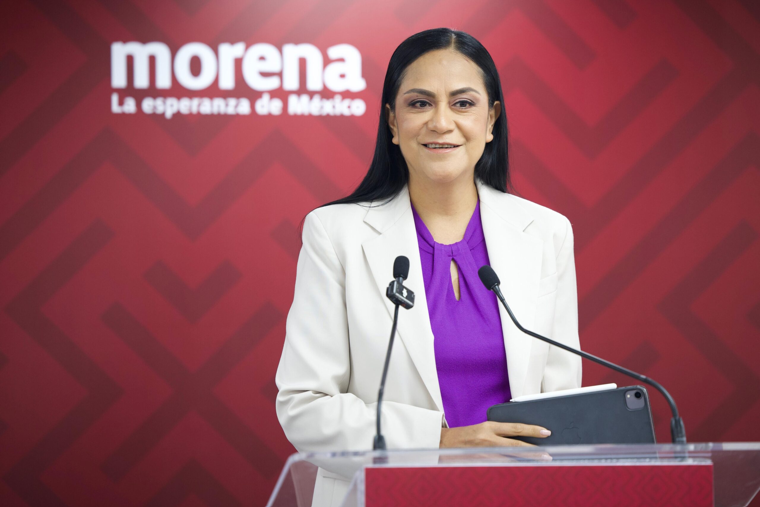 Mujer frente a dos micrófonos con el logotipo de Morena al fondo