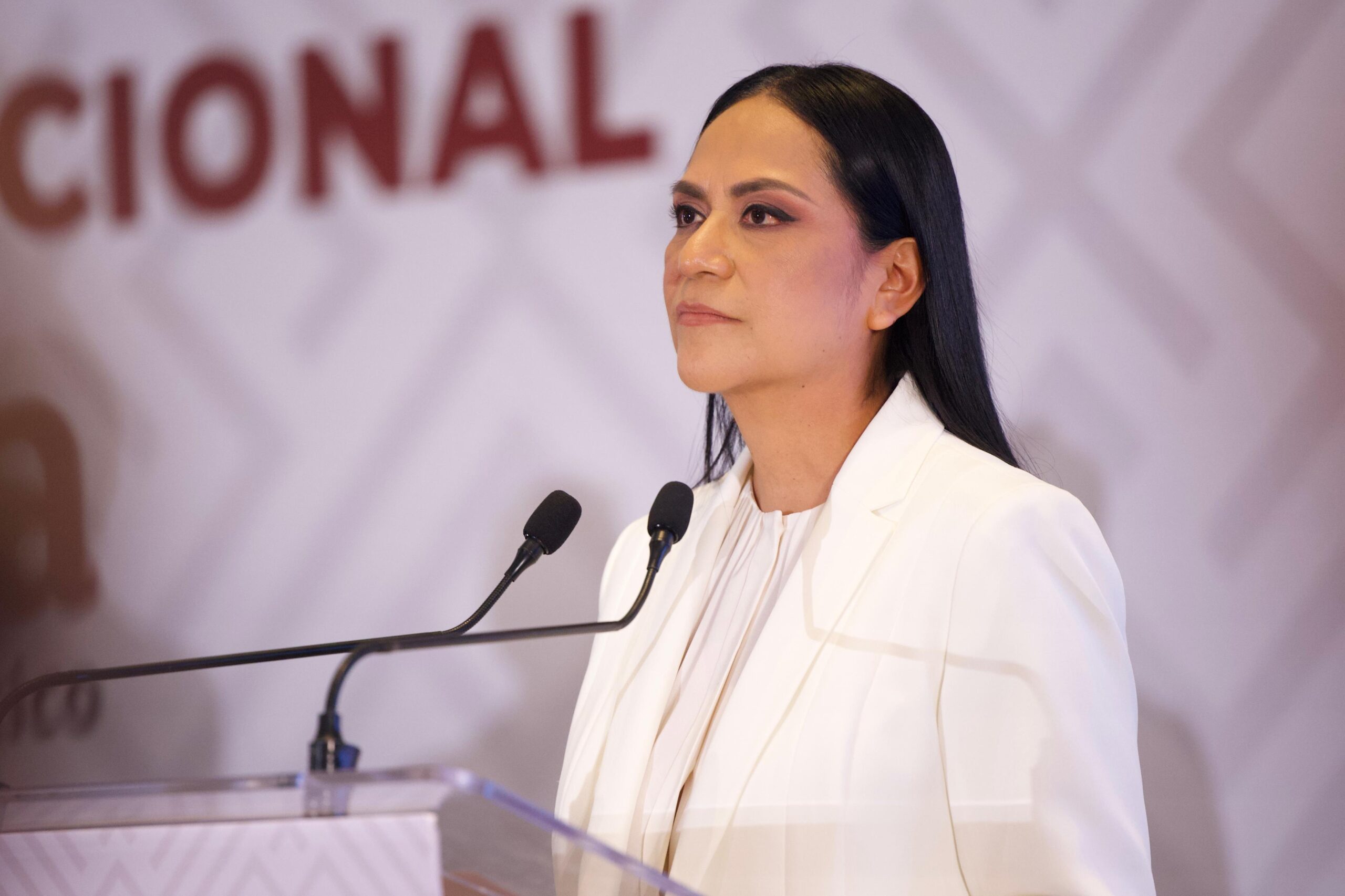 Mujer vestida de blanco frente a dos micrófonos durante un acto político