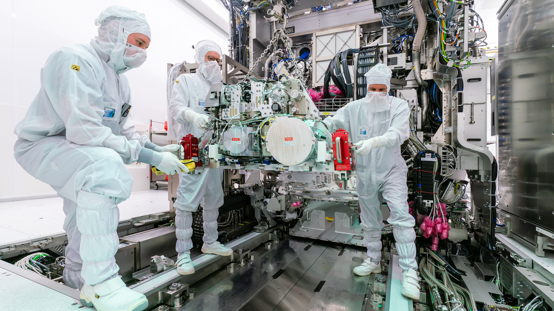 Tres trabajadores con trajes de sala limpia manipulan una pieza de maquinaria industrial de fabricación de semiconductores.