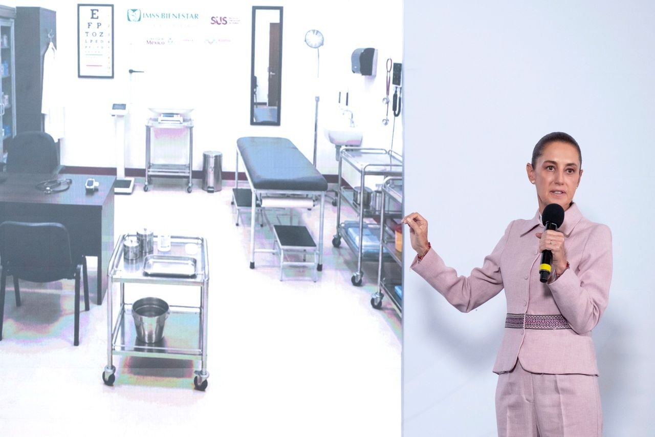 Mujer sostiene un micrófono durante una presentación frente a la imagen de un consultorio médico equipado