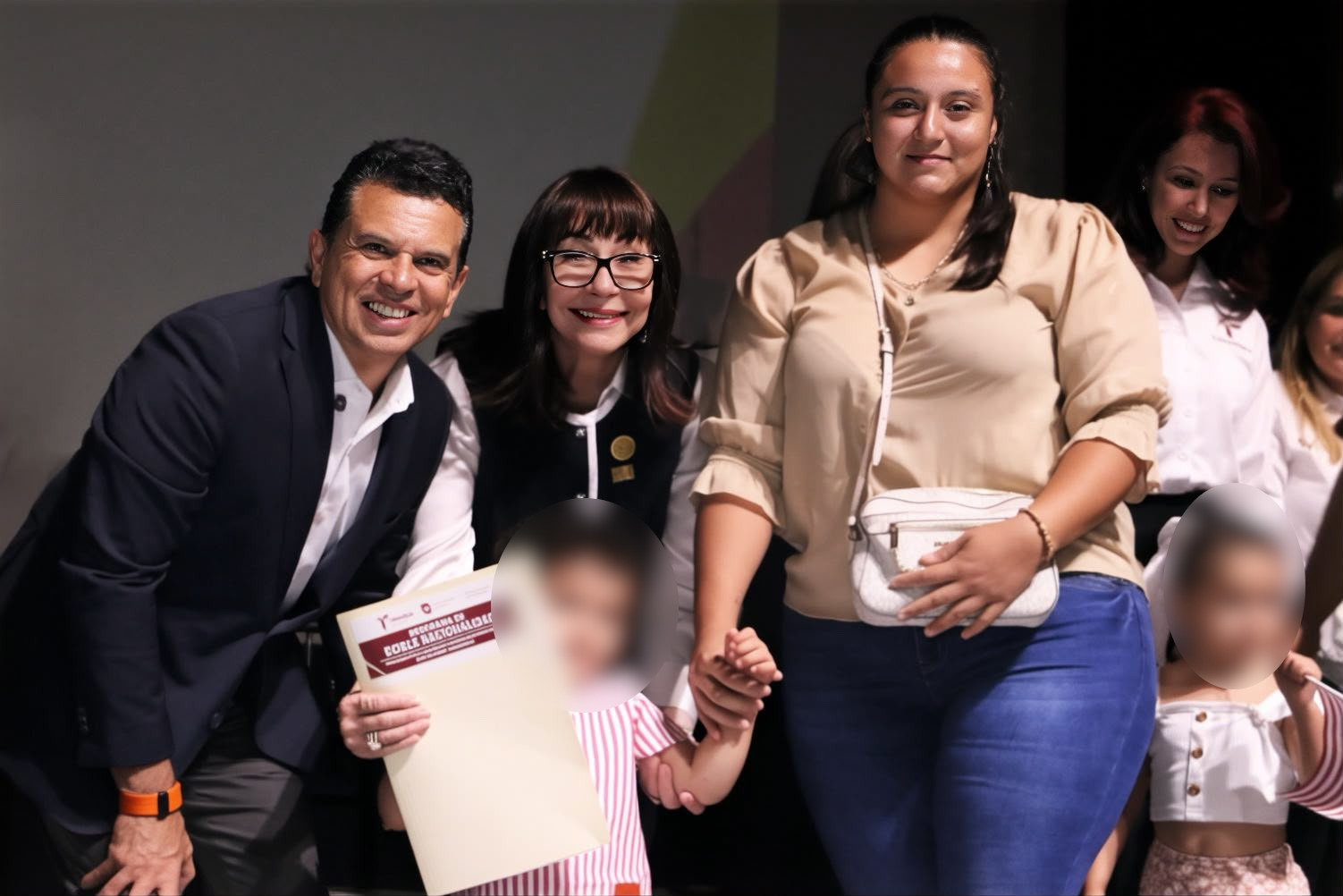Una mujer con lentes saluda a una niña pequeña durante un acto público