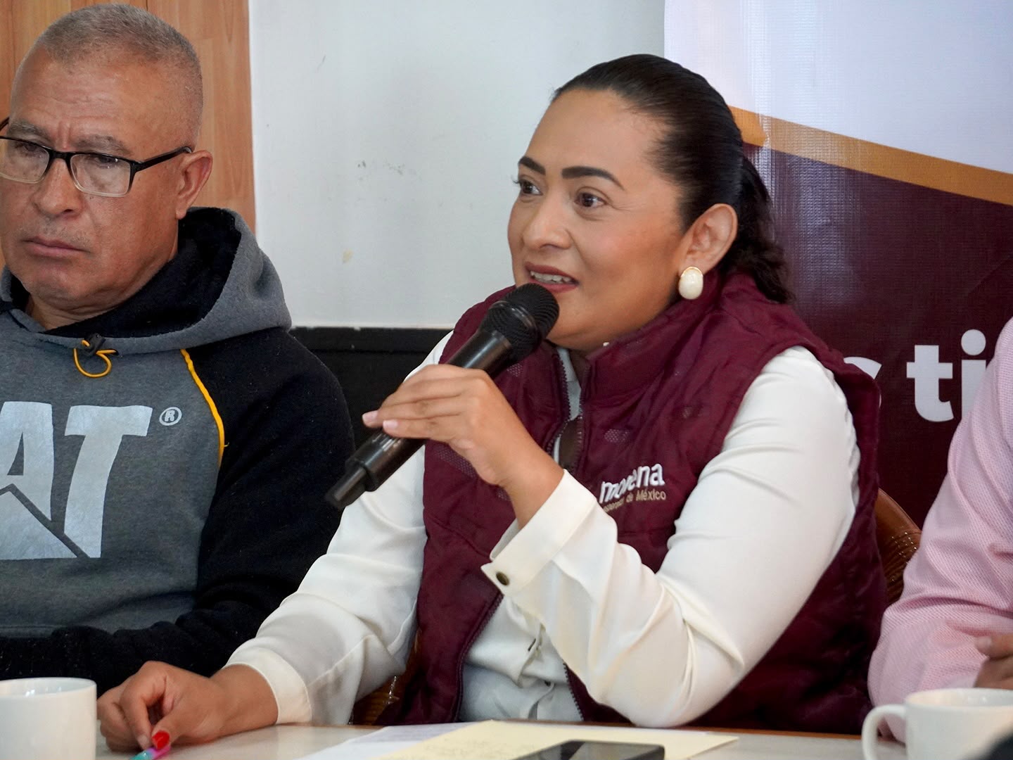 Una mujer con chaleco guinda de Morena habla por micrófono durante una reunión, junto a un hombre con lentes