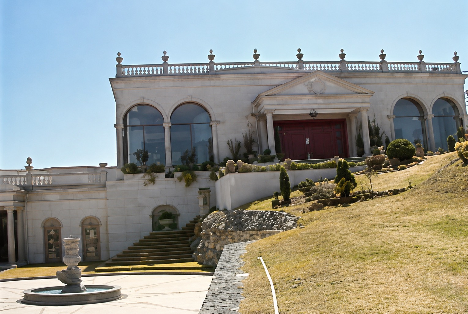 Fachada de piedra clara con ventanas arqueadas, grandes puertas rojas, escalinata y jardines en pendiente