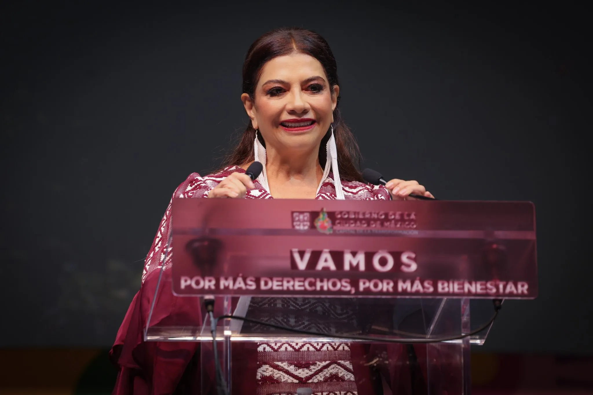 Mujer habla ante dos micrófonos desde un atril con la frase Vamos por más derechos, por más bienestar