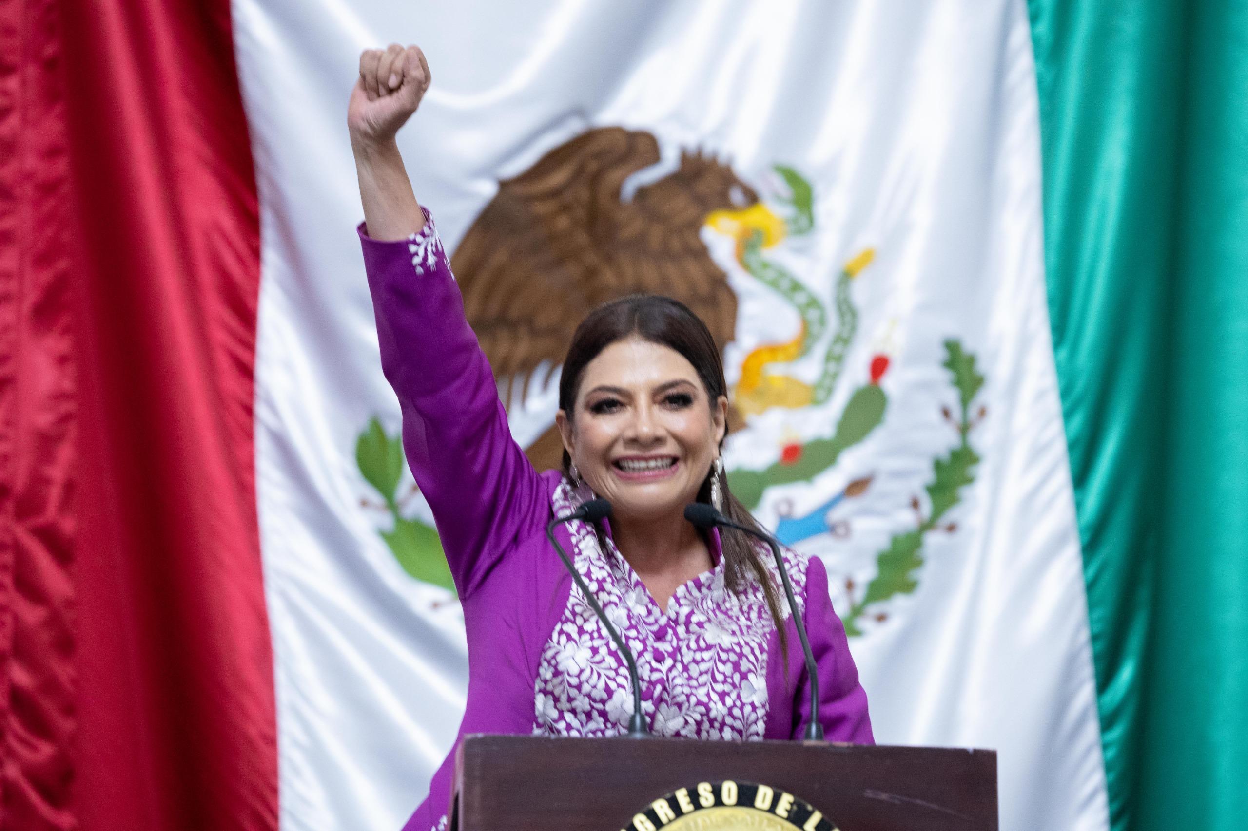 Clara Brugada durante su informe ante el Congreso de la Ciudad de México, con el puño levantado y la bandera nacional de fondo