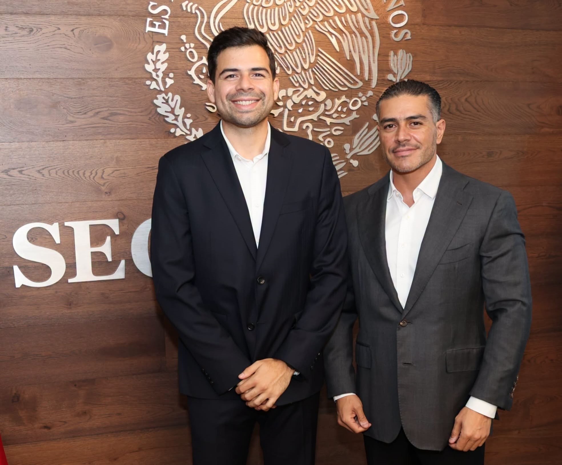 Manuel Cota Cárdenas y Omar García Harfuch posan frente al escudo nacional en una pared de madera