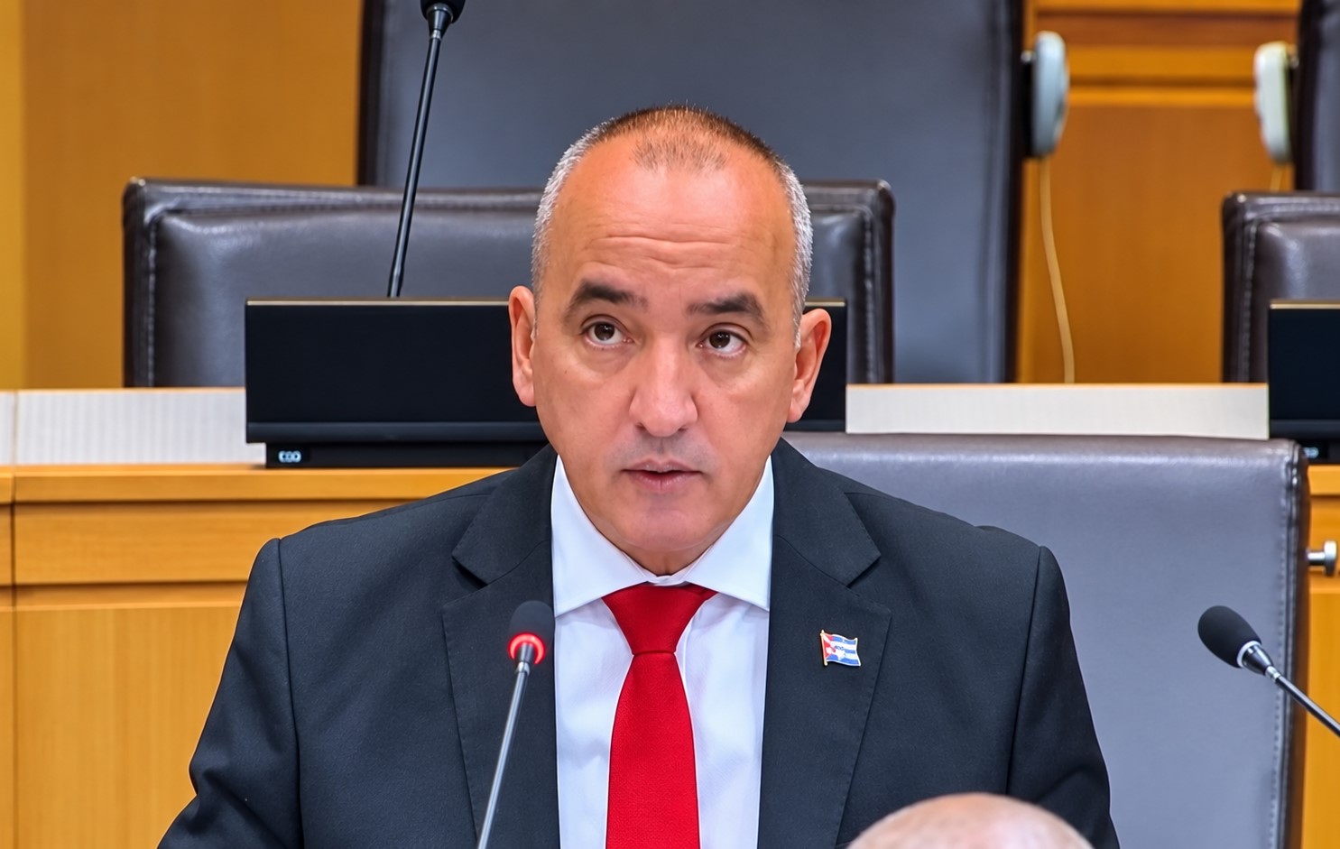 Diplomático de traje oscuro y corbata roja interviene ante micrófonos en una sala de reuniones.