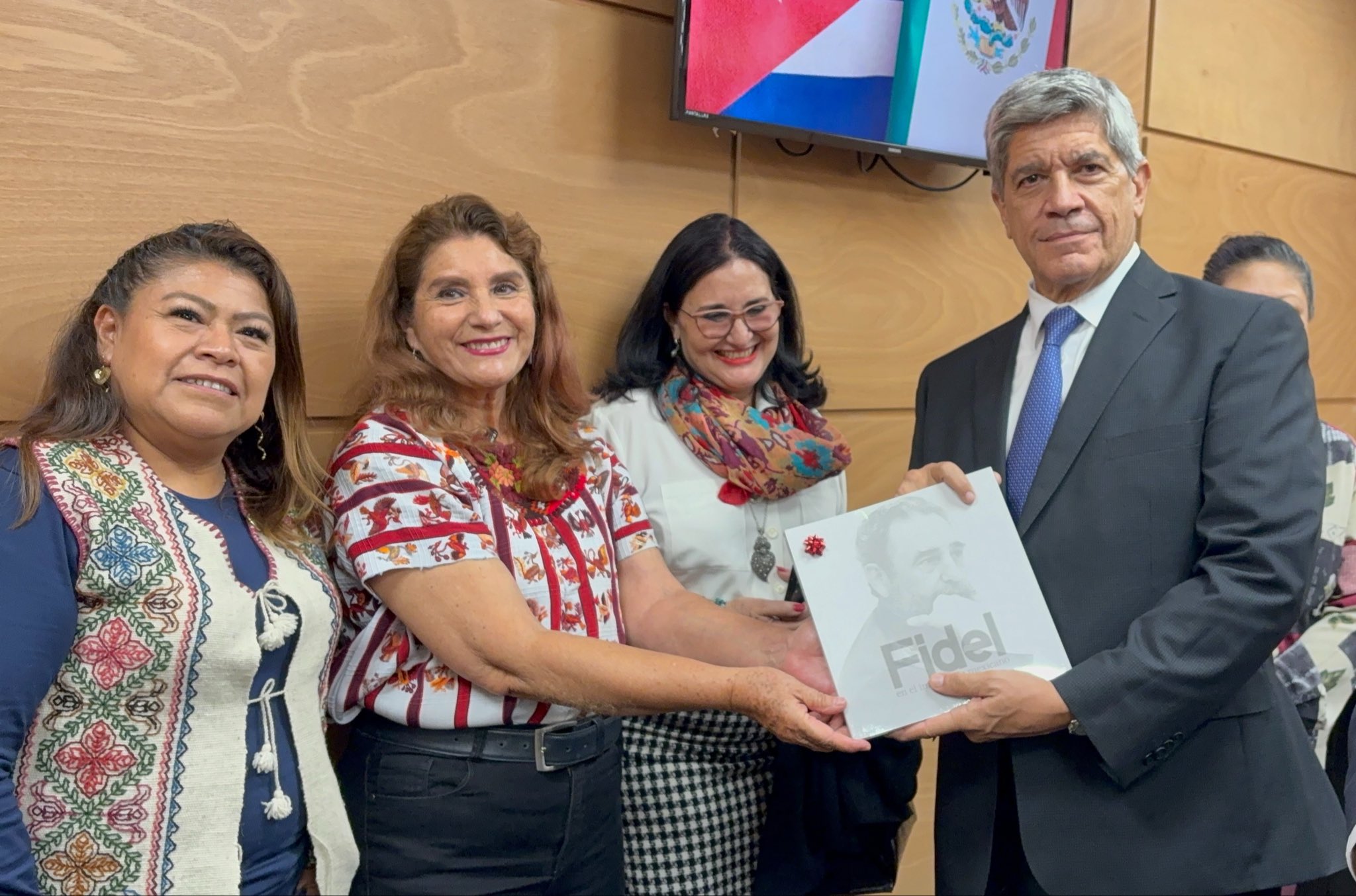 Cuatro personas posan durante un encuentro institucional mientras sostienen una publicación y al fondo se observan las banderas de México y Cuba
