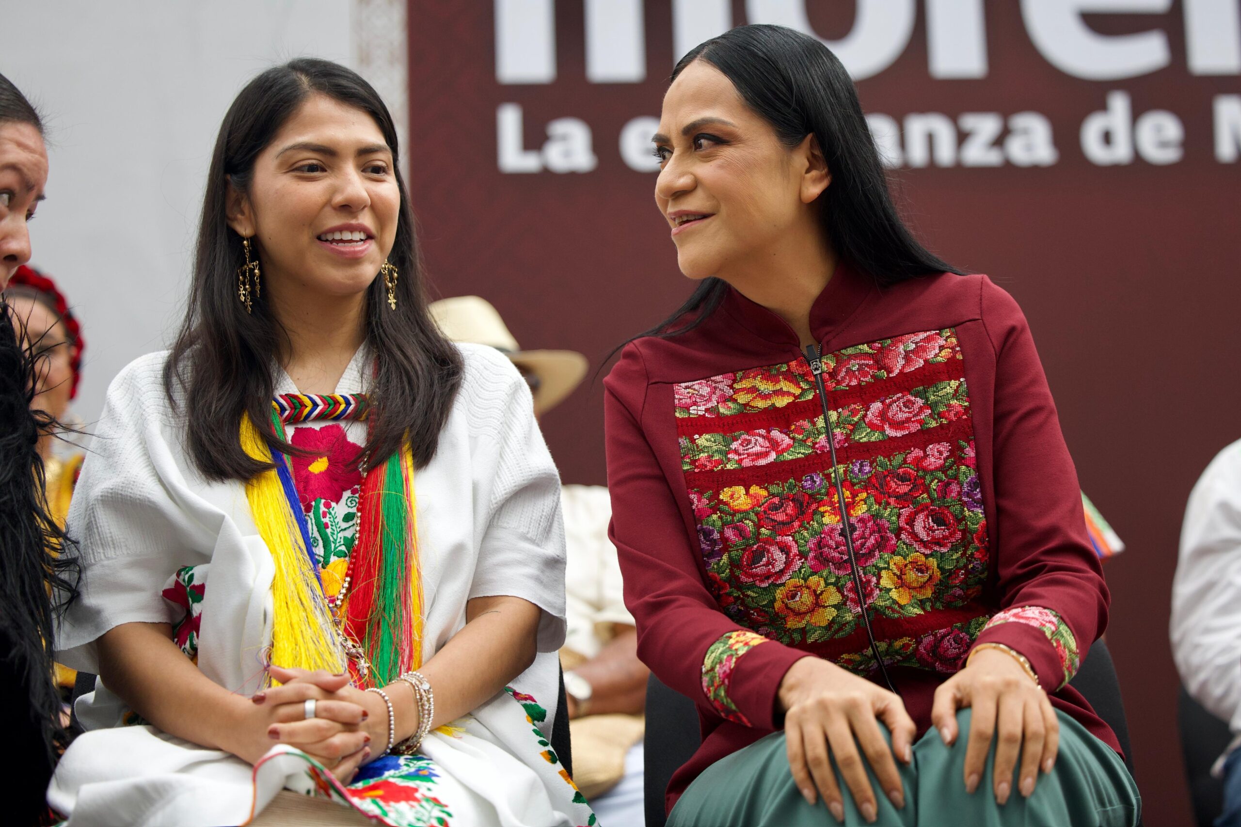 Dos mujeres con prendas bordadas conversan sentadas ante un fondo con el emblema de Morena durante un encuentro público