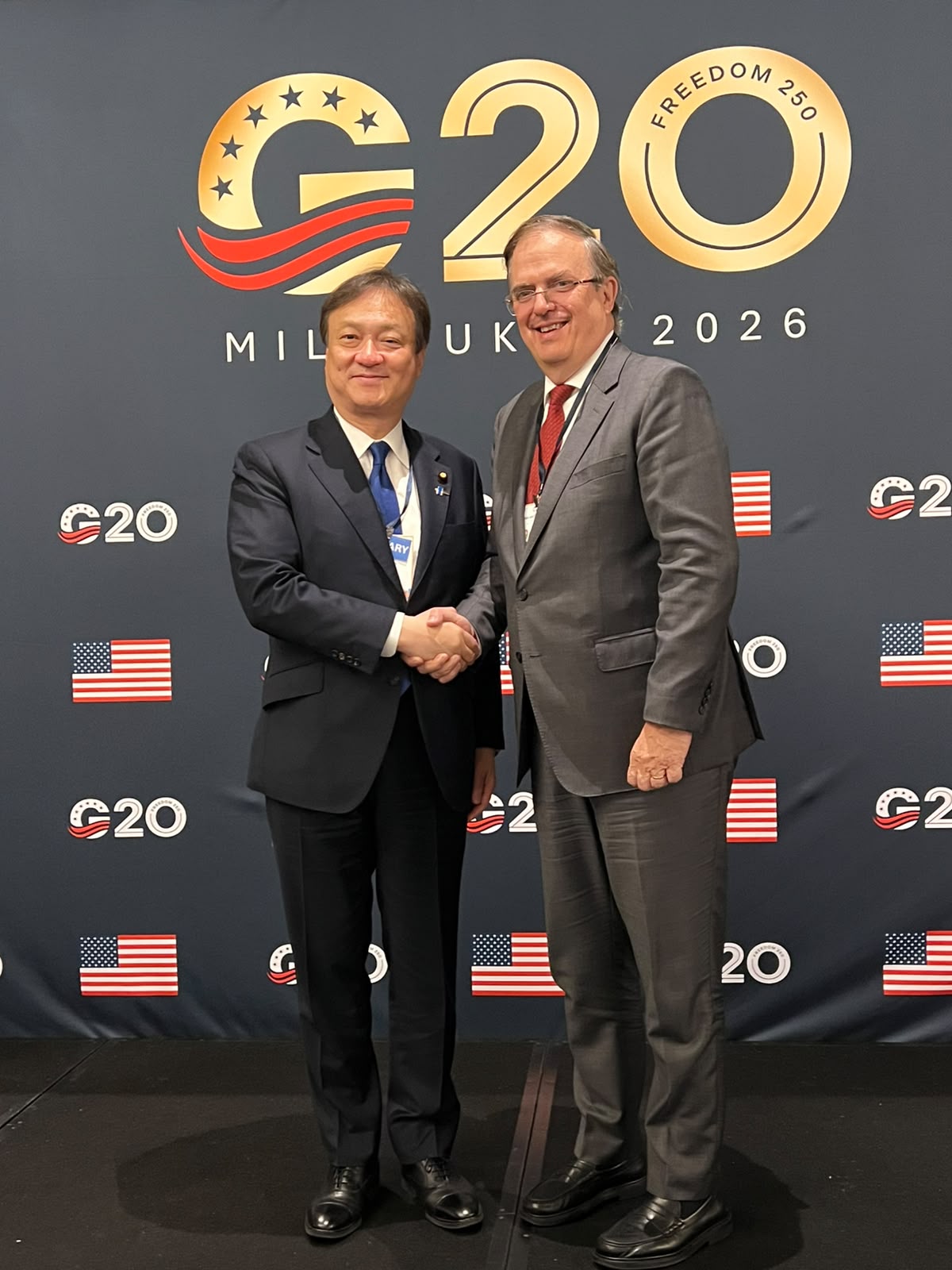 Marcelo Ebrard estrecha la mano de un funcionario japonés frente al emblema del G20 en Milwaukee