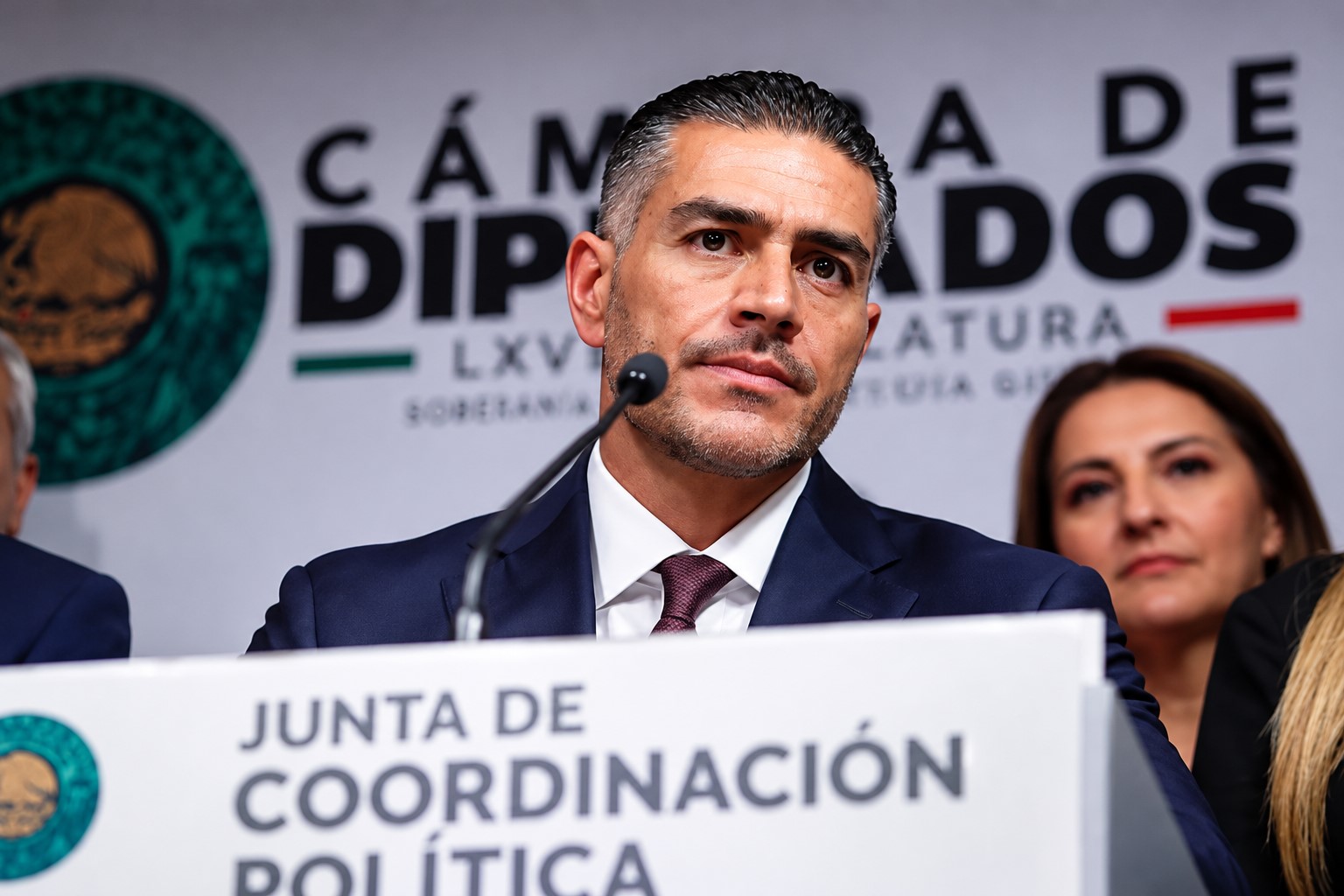 Un hombre de traje oscuro y corbata habla ante un micrófono durante un acto en la Cámara de Diputados; detrás aparecen otras personas y el emblema legislativo.