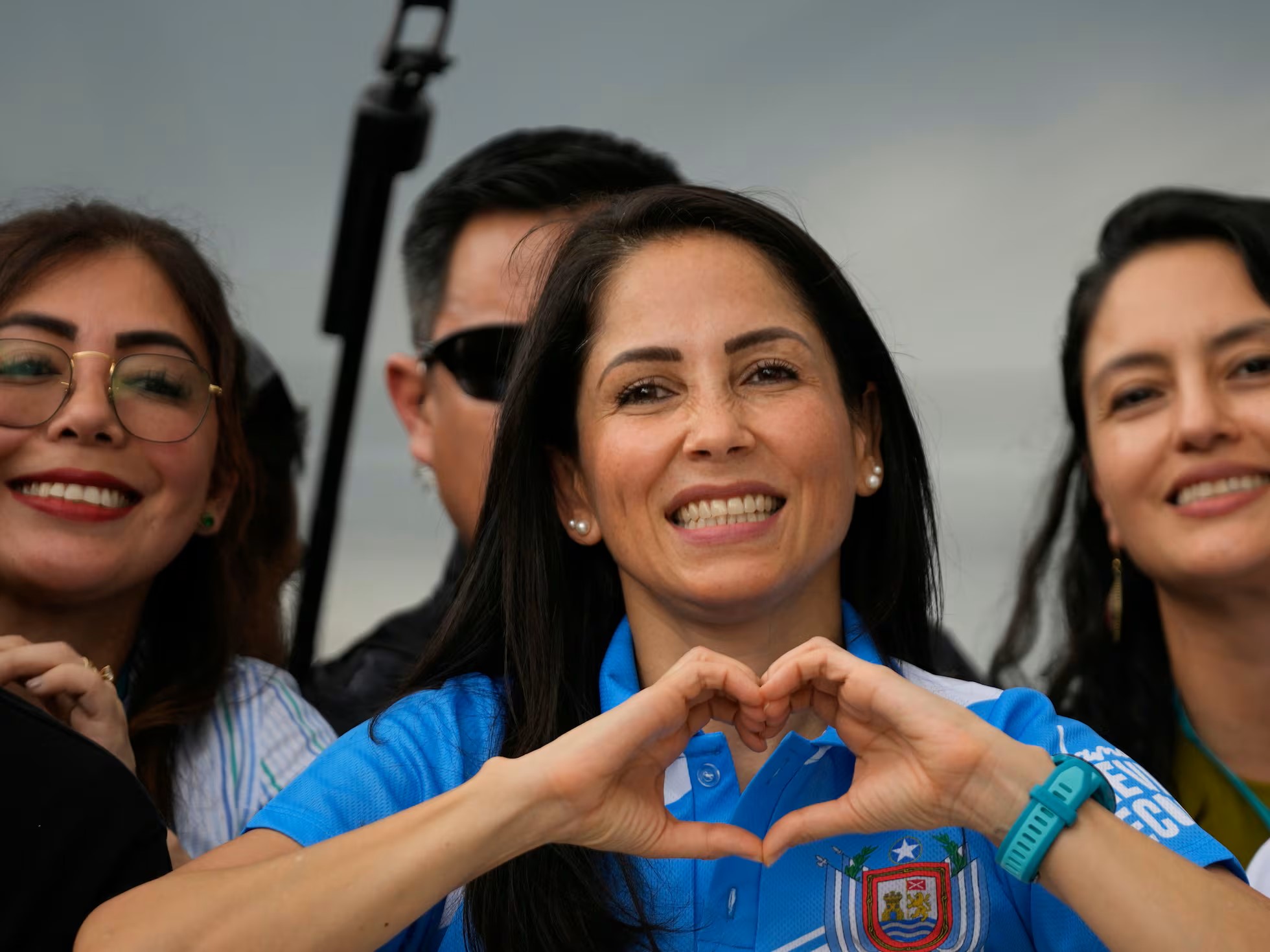 Luisa González sonríe y forma un corazón con sus manos durante una aparición pública, vestida de azul.