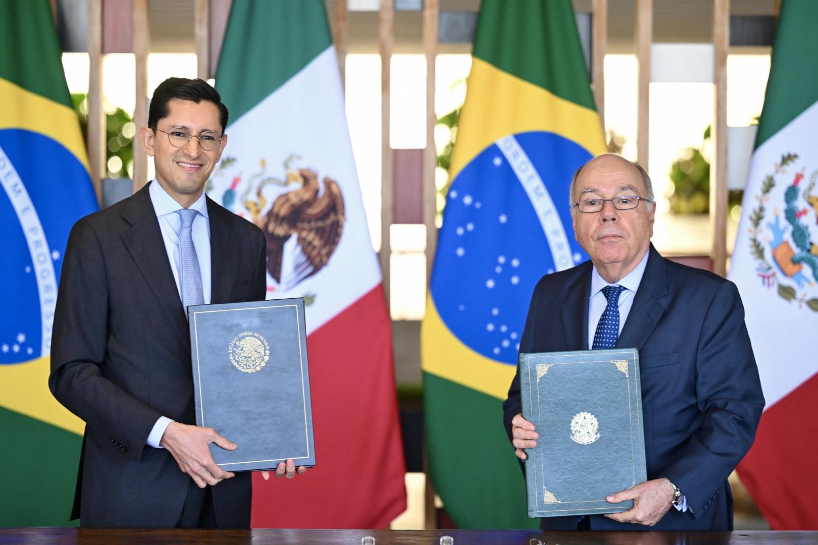 Dos funcionarios sostienen carpetas frente a banderas de México y Brasil