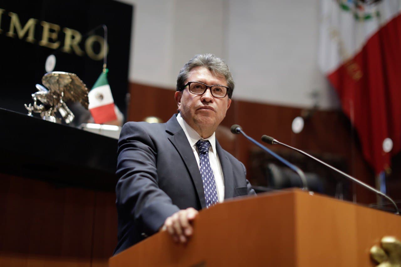 Legislador en la tribuna de la Cámara de Diputados, con micrófonos y la bandera de México al fondo