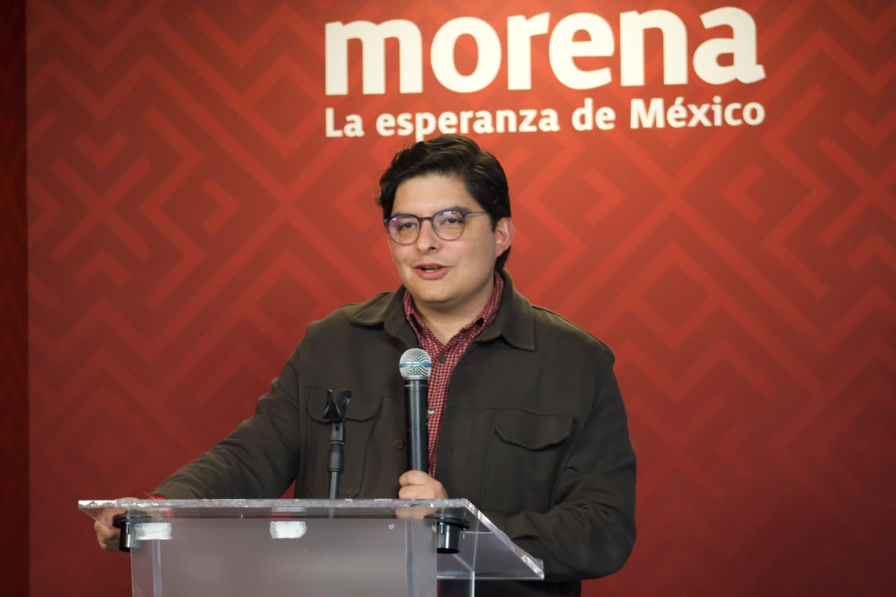 Hombre con lentes habla ante un micrófono y un atril, con el logotipo de Morena al fondo