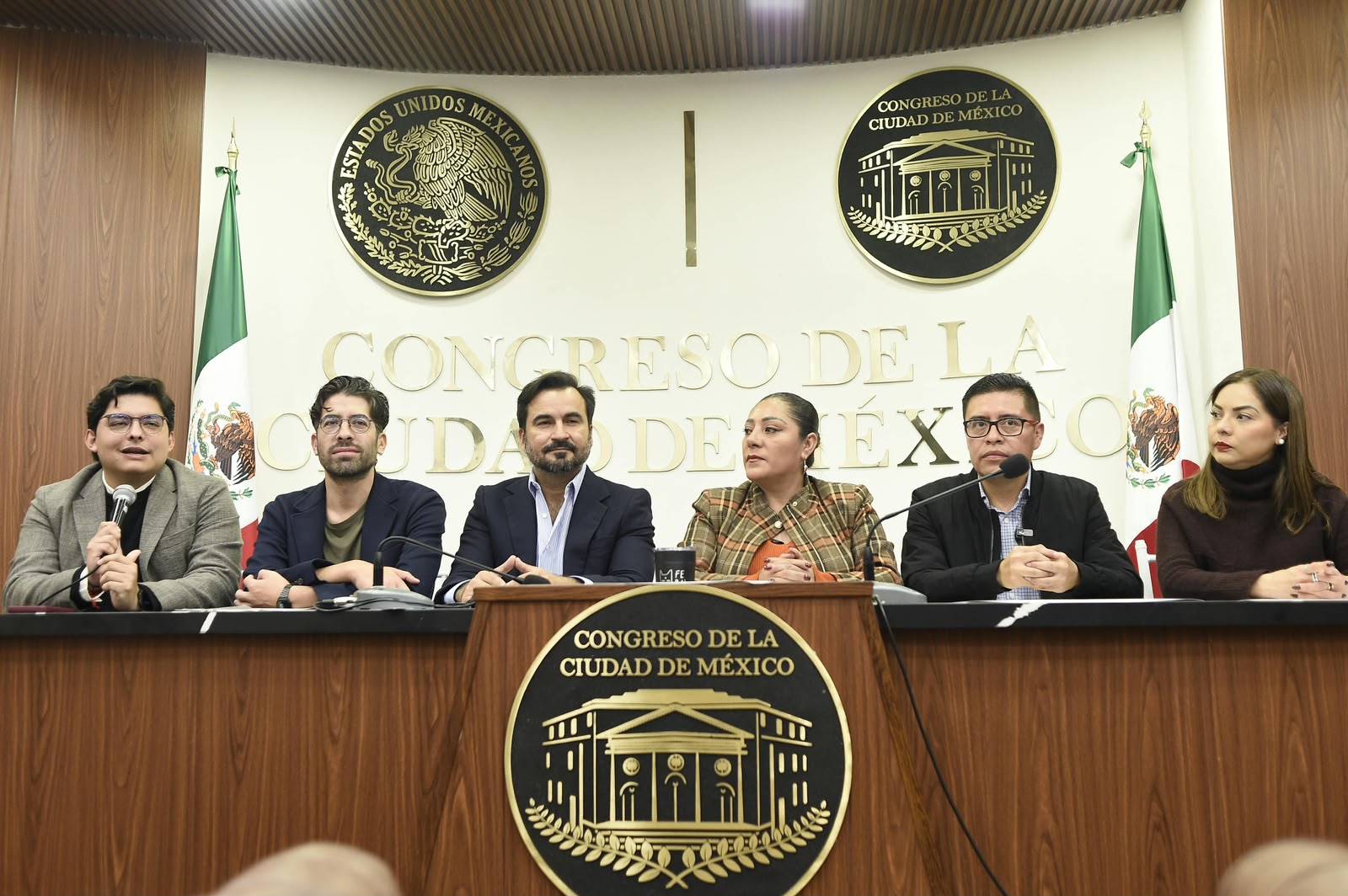 Seis participantes sentados ante micrófonos en una conferencia en el Congreso de la Ciudad de México.