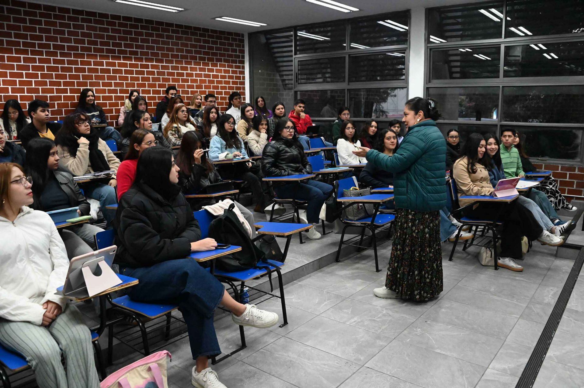 Grupo de estudiantes, en su mayoría mujeres jóvenes, escucha a una docente dentro de un aula