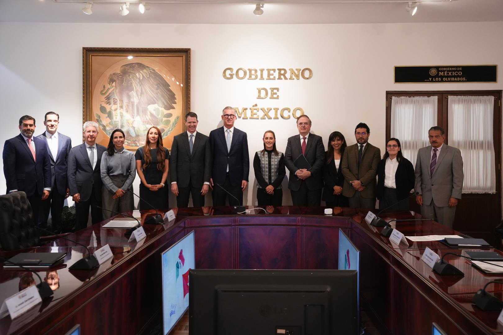 Grupo de personas posa en una sala de reuniones con el letrero Gobierno de México, una mesa de conferencias y un escudo nacional enmarcado.