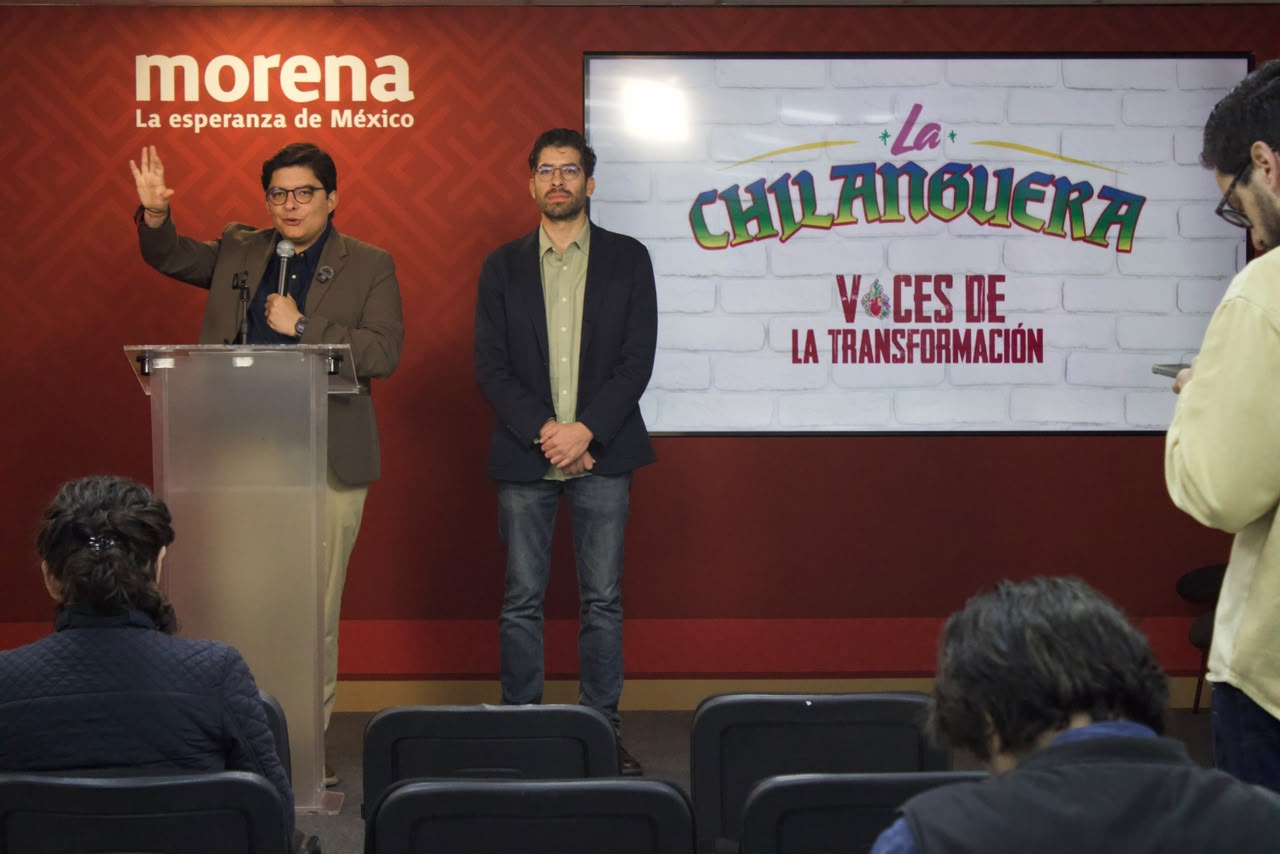 Conferencia de Morena con un orador frente al micrófono y otra persona junto a una pantalla de La Chilanguera