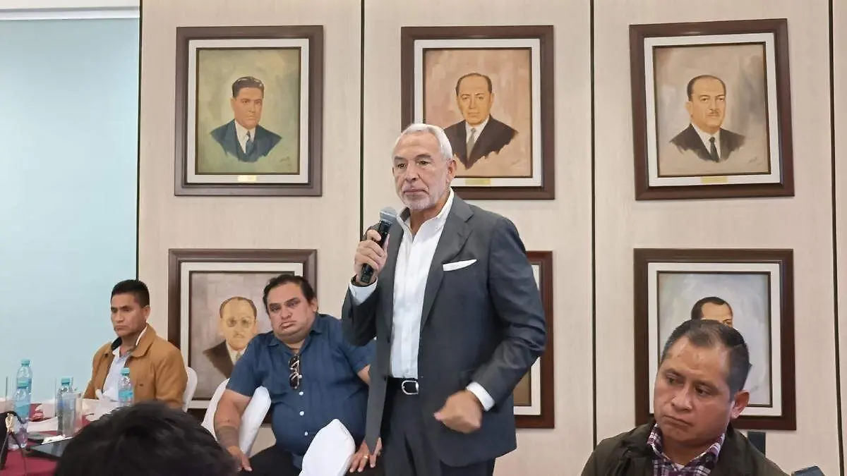 Hombre con traje sostiene un micrófono durante una reunión en un salón con varias personas sentadas