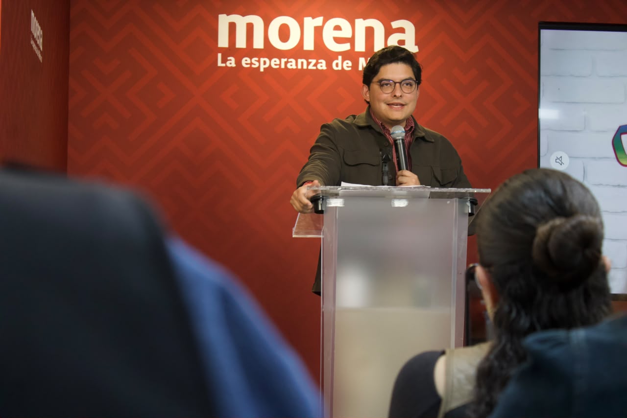 Un hombre con lentes habla ante un micrófono detrás de un atril transparente, frente a un fondo rojo con el logotipo de Morena y asistentes en primer plano