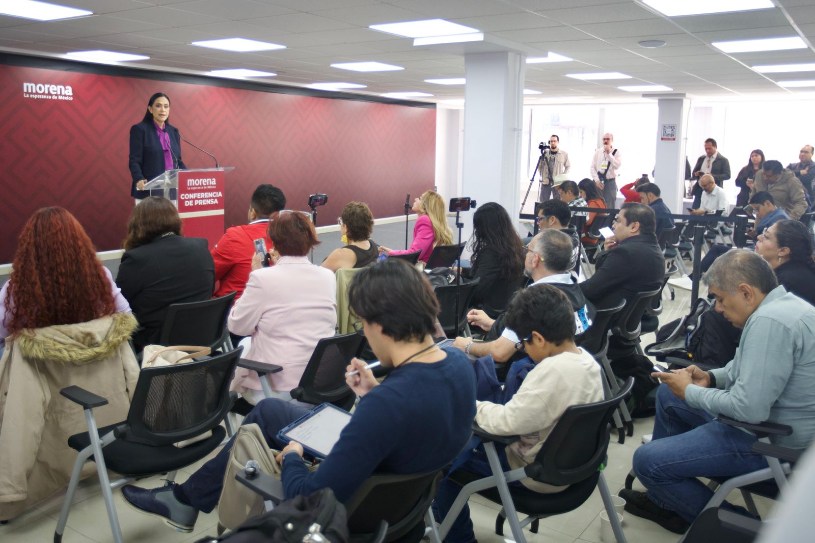 Conferencia de prensa de Morena con una oradora frente a asistentes y periodistas en una sala