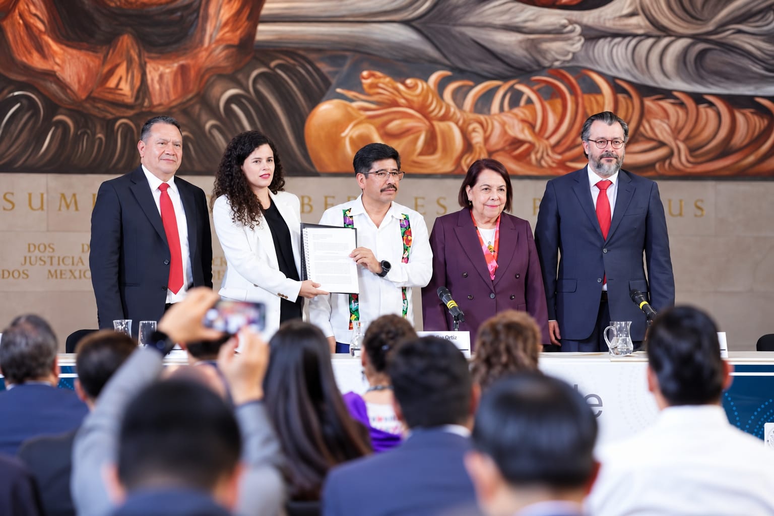 Dos participantes en una conferencia institucional frente a micrófonos y documentos